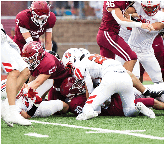 Several football players in helmets and uniforms are engaged in a tackle on a field with yard lines visible. Description generated by AI