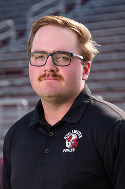 A person with short hair, glasses, and a mustache is wearing a black polo shirt with a Stillwater Ponies logo and standing in front of blurred stadium bleachers. Description generated by AI