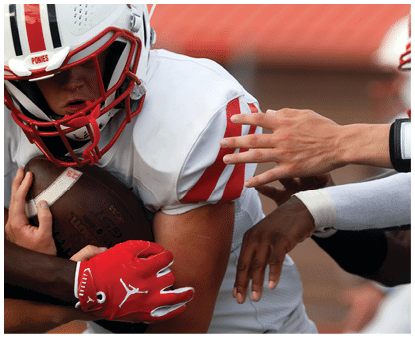 A person in a white football uniform with red accents is holding a football tightly while another person reaches out to tackle or block them. Description generated by AI