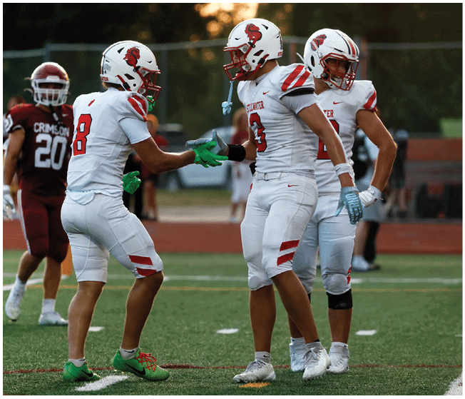 Three people in white football uniforms are on a field, with two of them shaking hands while a person in a maroon uniform stands in the background. Description generated by AI