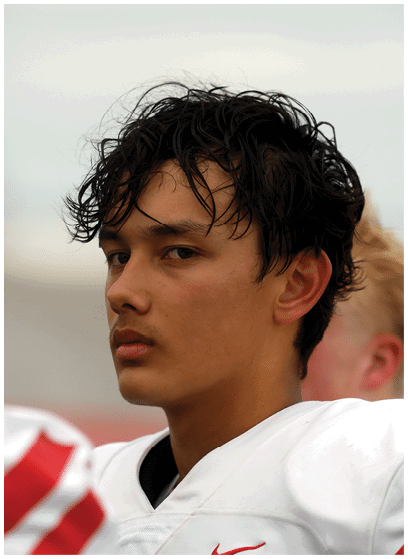 A person with wet, tousled hair wearing a white sports jersey looks thoughtfully to the side against a blurred outdoor background. Description generated by AI