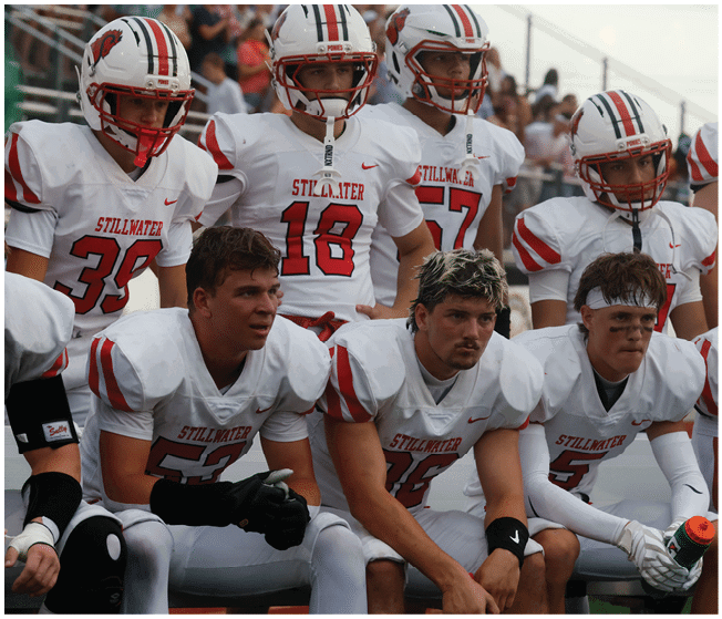 A group of people wearing white football uniforms with red and black accents are sitting and standing closely together on a sports field. Description generated by AI