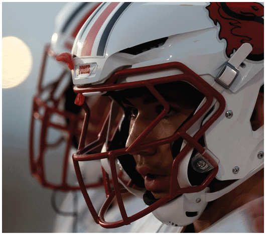 Two people wearing white football helmets with red faceguards and a red lion logo are focused and looking ahead. Description generated by AI