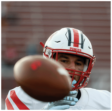 A person wearing a white football helmet with red and black stripes and a white jersey is catching or throwing a football close to the camera. Description generated by AI
