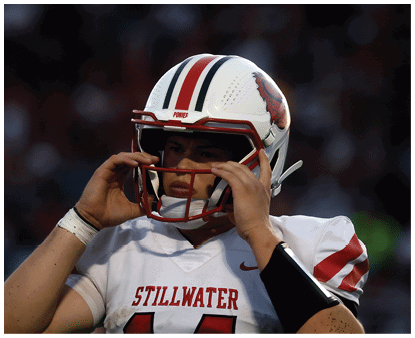 A person wearing a football helmet and a white jersey with “Stillwater" written on it is adjusting their helmet on a field with a blurred crowd in the background. Description generated by AI