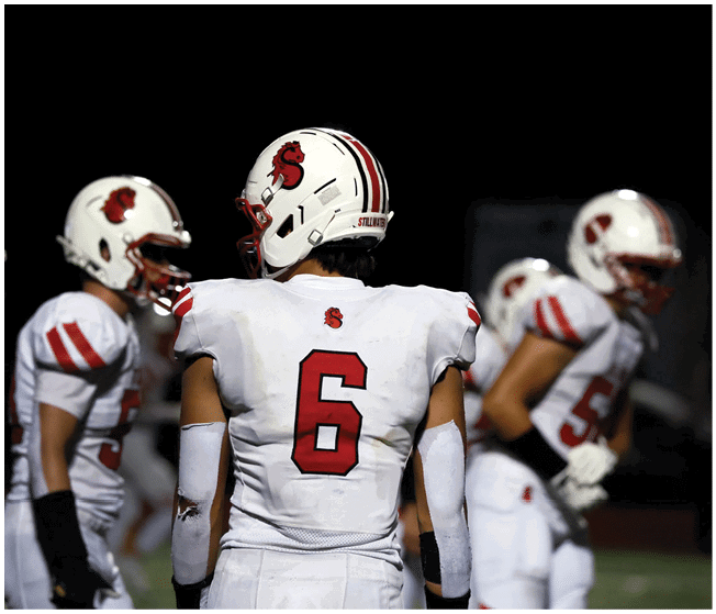 Four people in white football uniforms with red accents stand on a field at night, with one person facing away showing the number six on their jersey. Description generated by AI