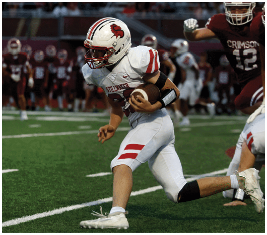 A person in a white football uniform with red stripes is running with a football on a green field while players in maroon uniforms are in the background. Description generated by AI