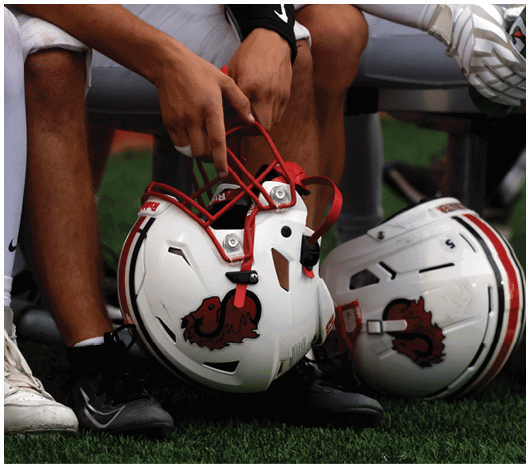 Two people wearing athletic shoes and sports gear are sitting on a bench, one holding a white football helmet with red accents and a red lion logo. Description generated by AI