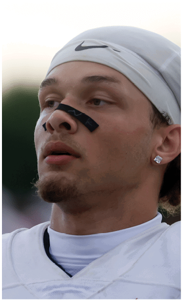 Person wearing a white sports head covering and jersey with black athletic tape across the nose looking slightly upward. Description generated by AI
