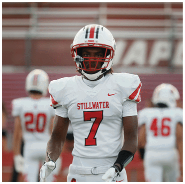 A person wearing a white football uniform with the number seven and the word Stillwater stands in the foreground on a football field while another person in a similar uniform with the number 46 is blurred in the background. Description generated by AI