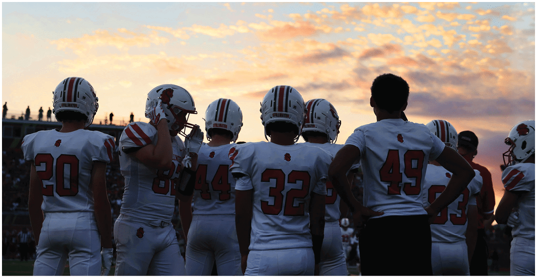 A group of people wearing football uniforms with helmets stand together on a field at sunset. Description generated by AI