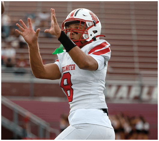 A person wearing a white football uniform with the number 8 and a helmet is reaching up with both hands as if to catch a ball on a football field. Description generated by AI