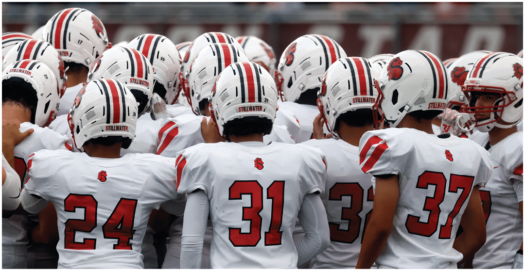 A group of people wearing white football uniforms with red numbers and helmets are huddled together on a field. Description generated by AI
