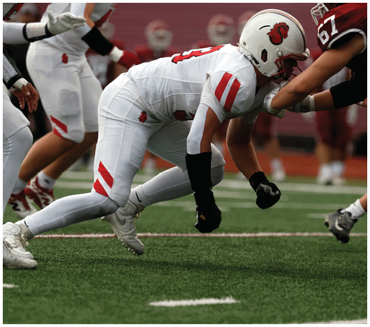 A person in white football gear with red accents is crouched low on a green field, engaging with another player in maroon gear during a game. Description generated by AI