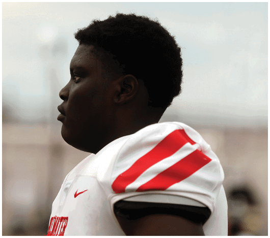 A person wearing a white sports jersey with red stripes on the shoulder is shown in profile against a blurred outdoor background. Description generated by AI