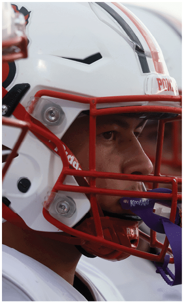 People wearing white football helmets with red faceguards stand closely together, focused on the game. Description generated by AI