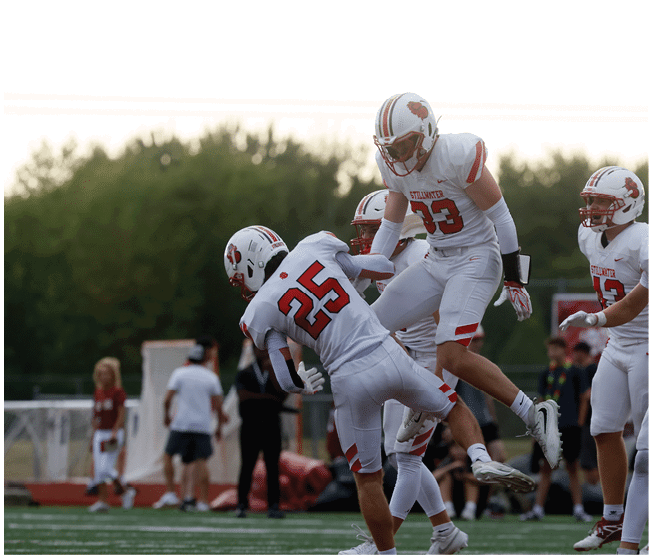 Three people in white football uniforms with red numbers are celebrating on a football field while others stand in the background. Description generated by AI
