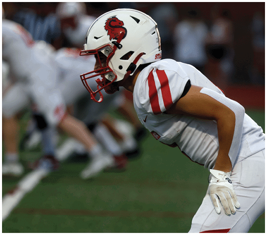 A person in a white football uniform with red stripes and a helmet is leaning forward in a ready position on a football field. Description generated by AI
