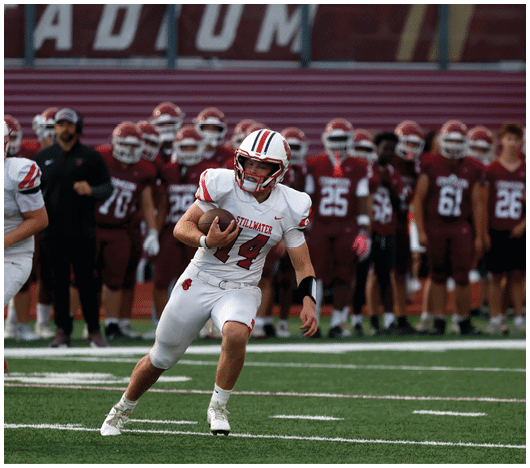 A person in a white football uniform with the number 14 is running with the ball on a football field while a group of people in maroon uniforms stand in the background. Description generated by AI