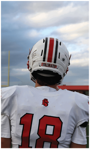 A person wearing a white football uniform with the number 18 and a helmet labeled “STILLWATER" stands on a football field facing away from the camera under a cloudy sky. Description generated by AI