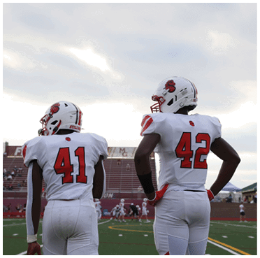 Two people in white football uniforms with red numbers stand on a football field with a stadium and other players in the background under a cloudy sky. Description generated by AI