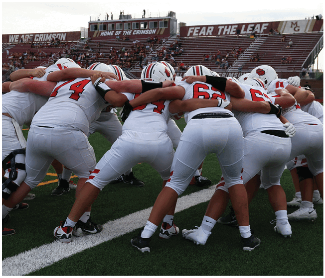 A group of people in white football uniforms with red and black accents are huddled together on a football field with empty bleachers in the background. Description generated by AI