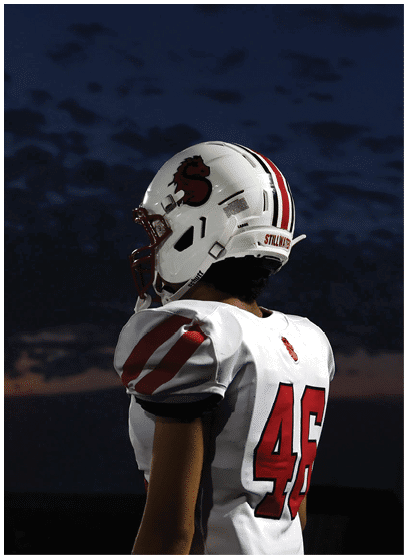A person wearing a white football helmet and uniform with red stripes is standing against a darkening sky at sunset. Description generated by AI