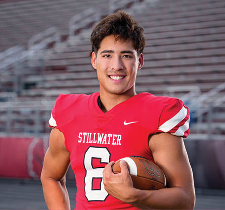 A person wearing a red football jersey with the number 6 and the word Stillwater is smiling and holding a football in a stadium. Description generated by AI