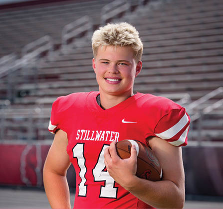 A person with short blond hair wearing a red football jersey with the number 14 and the word Stillwater is smiling while holding a football in a stadium. Description generated by AI