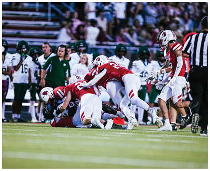 Several people in red and white football uniforms are tackling a person in a dark uniform on a football field while a referee and others watch in the background. Description generated by AI