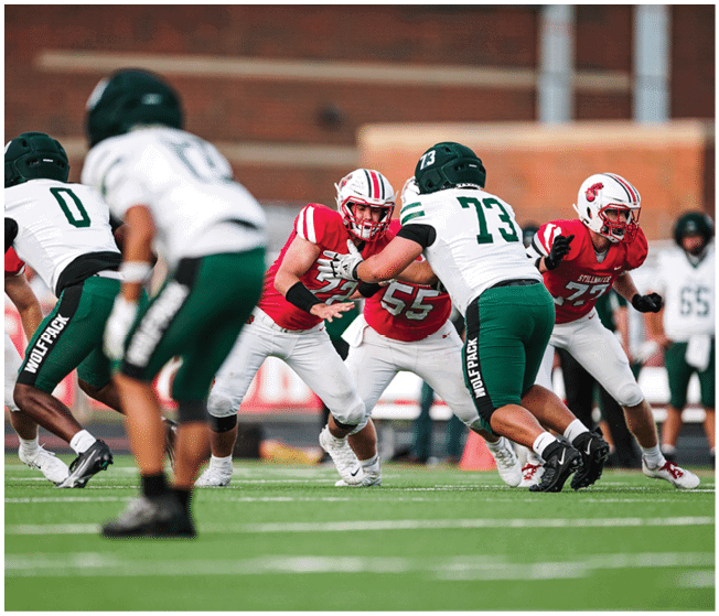 Players in red and white uniforms and players in white and green uniforms are engaged in a football game on a grassy field. Description generated by AI