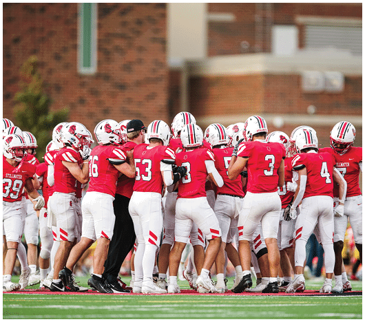 A group of people wearing red and white football uniforms are huddled together on a field during a game. Description generated by AI