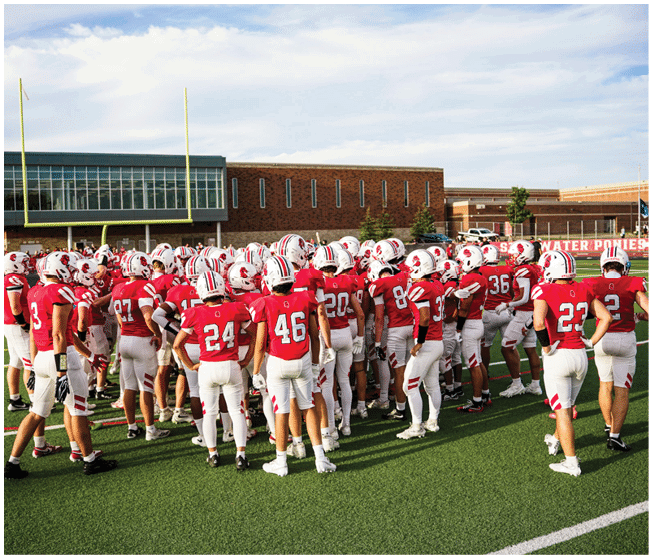 A group of people wearing red and white football uniforms and helmets are gathered on a green football field near goalposts and a school building. Description generated by AI