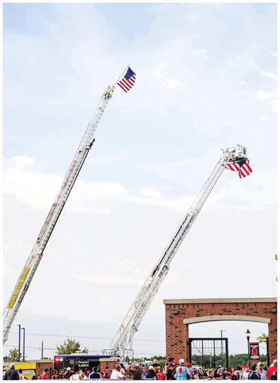 Two fire truck ladders extended high with American flags attached at the top, above a crowd of people near a brick archway under a partly cloudy sky. Description generated by AI