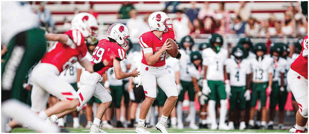 Adults in football uniforms are actively playing on a field with one person holding a football and others running. Description generated by AI