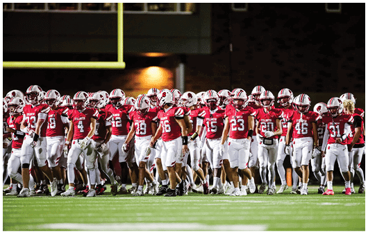 A large group of people wearing red and white football uniforms are gathered on a green field under stadium lights at night. Description generated by AI