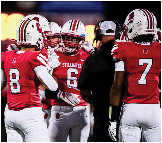 People in red and white football uniforms are gathered on a field, with one person wearing a headset and others appearing to communicate during a game. Description generated by AI