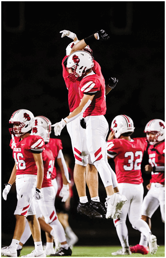 Two people in red and white football uniforms with helmets are raising their arms during a game, with others in similar uniforms visible in the background. Description generated by AI