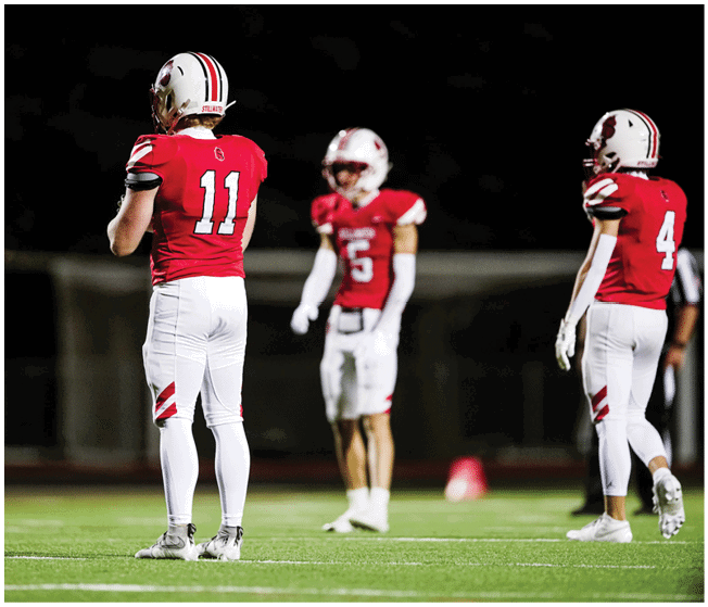 Three people wearing red and white football uniforms stand on a field at night, with one person holding a football. Description generated by AI