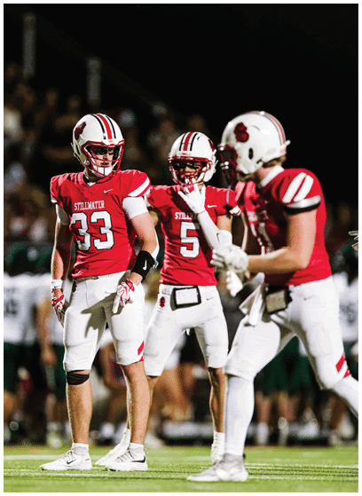 Three people in red and white football uniforms stand on a field during a nighttime game, with one person in the foreground and two others in the background. Description generated by AI