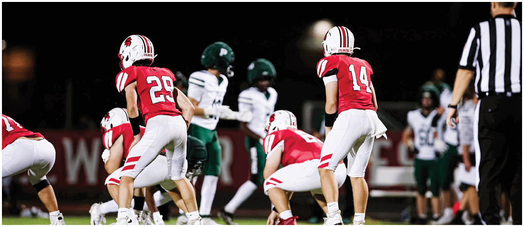 People in red and white football uniforms are positioned on a field facing opponents in green and white uniforms during a nighttime game. Description generated by AI