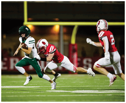 A person in green and white sports gear is running with a football while another person in red and white sports gear lunges to tackle them on a football field at night. Description generated by AI