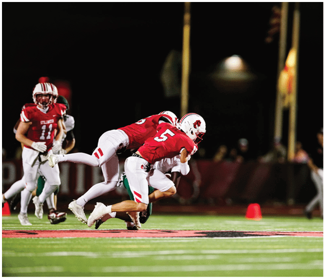 Two people in red and white football uniforms are engaged in a tackle on a lit football field at night. Description generated by AI