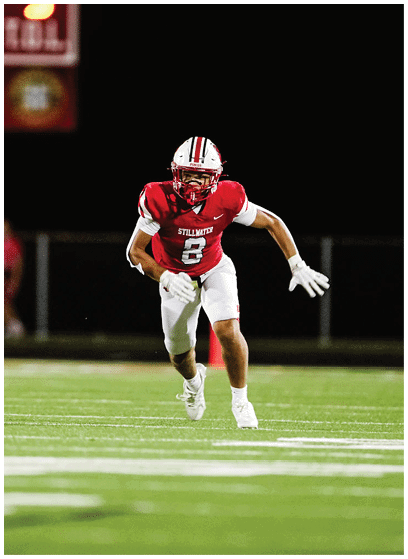 A person in a red and white football uniform is running on a field toward another person in a green and white uniform with a scoreboard visible in the background. Description generated by AI