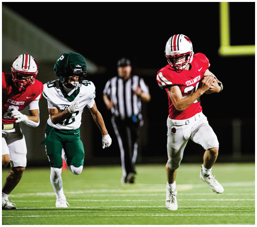 A person in a red football uniform is running with the ball while being chased by a person in a white and green uniform on a football field at night. Description generated by AI