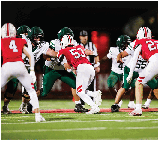 Players in red and white uniforms and players in green and white uniforms are engaged in a football game on a field at night. Description generated by AI