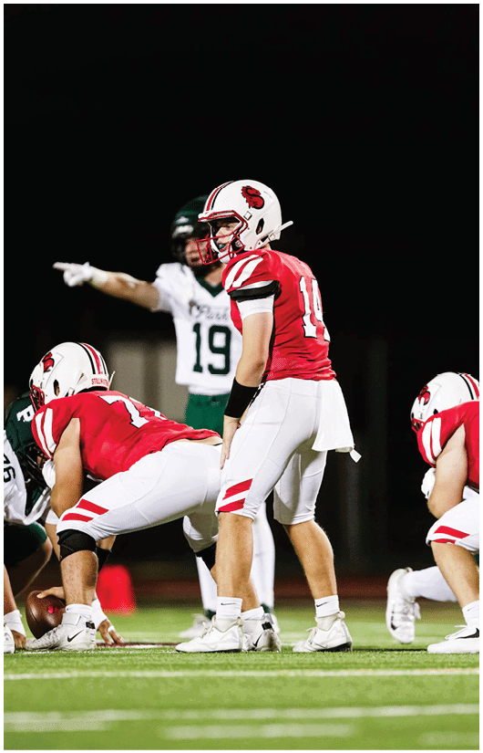 People in football uniforms are positioned on a field at night, with one person in a red jersey standing behind the line of scrimmage and others crouched in preparation for a play. Description generated by AI