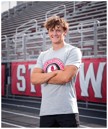 A person with tousled hair wearing a gray t-shirt with a sports logo stands smiling with arms crossed on a track field in front of bleachers and a red banner. Description generated by AI