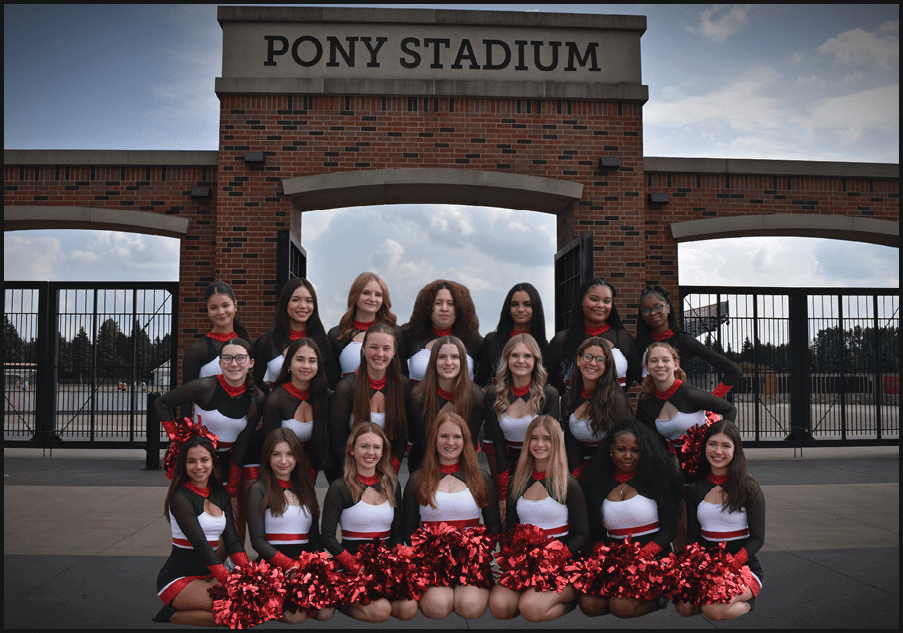 A group of adults dressed in matching cheerleading uniforms with red and black colors pose together outdoors holding red pom-poms in front of a brick building and metal fence. Description generated by AI