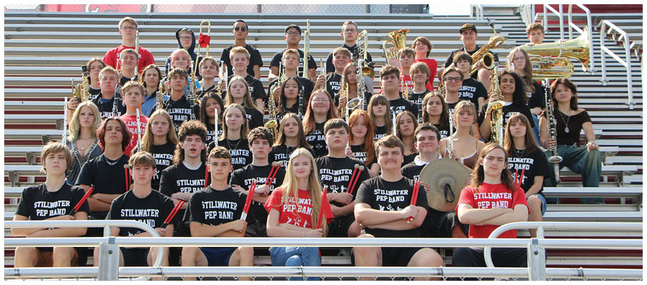 A large group of people wearing matching Stillwater Pep Band shirts are seated in bleachers holding musical instruments. Description generated by AI